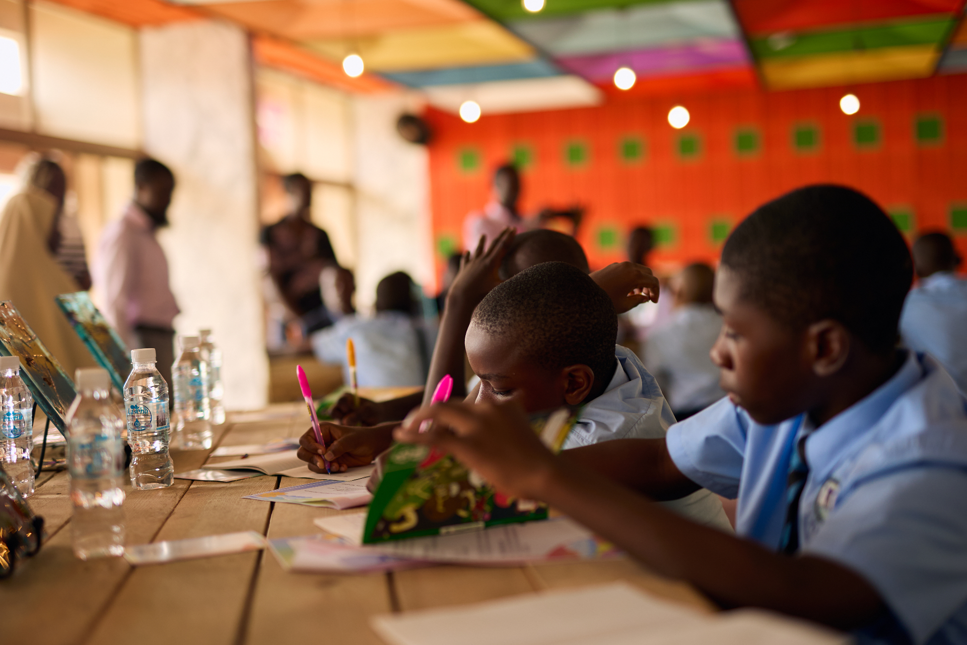 Children writing at a long wooden table in a brightly coloured workshop space